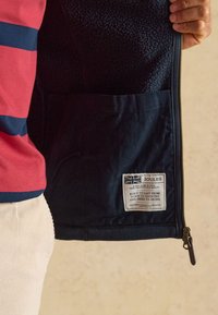 Hand hält eine offene marineblaue Jacke, die das Fleecefutter, eine Innentasche, ein Etikett mit britischer Flagge und Text über Qualität und Handwerkskunst zeigt.