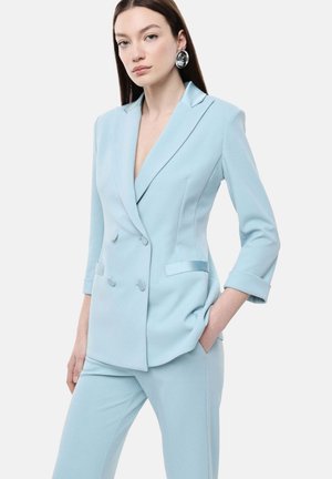 Donna che indossa un blazer doppiopetto azzurro chiaro con maniche arrotolate e pantaloni abbinati, posando con una mano in tasca.