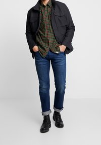 Chaqueta negra con dos bolsillos en el pecho, camisa de cuadros verde y roja, jeans azul oscuro arremangados en los puños, botas negras con cordones.