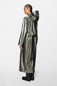 Μακριά μεταλλική πράσινη αδιάβροχη με κουκούλα, με εφαρμοστό στη μέση και χαλαρή γραμμή. Συνδυάζεται με μαύρες λαστιχένιες μπότες.