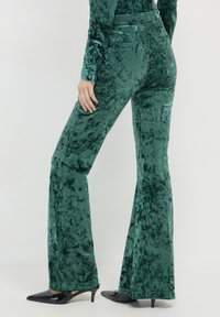 Pantaloni a zampa d'elefante in velluto verde con una texture liscia, dotati di vita media e una leggera lucentezza, abbinati a tacchi a punta neri.
