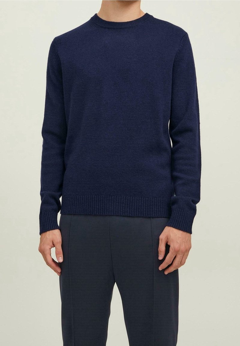 Uomo che indossa un maglione a girocollo blu navy a maniche lunghe e pantaloni scuri, in piedi davanti a uno sfondo chiaro.