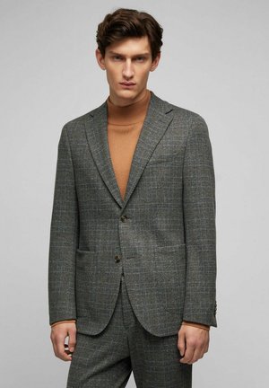 Grau kariertes Blazer mit Revers, zwei-Knopf-Verschluss und aufgesetzten Taschen, kombiniert mit einem braunen Rollkragenpullover darunter.