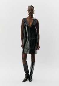 Must mustrüü mustast nahast, V-kaeluse, ilma varrukateta disainiga, kahe ees oleva tasku ja sirge lõikega. Mudel kannab kuni põlveni ulatuvaid musti saapaid.