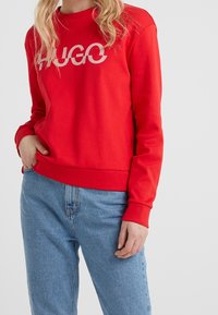 Sudadera roja de manga larga y cuello redondo, con un gran logotipo blanco "HUGO" en la parte frontal. El material parece suave y con textura.