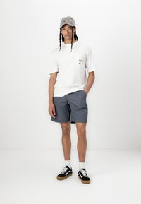 Hvit T-skjorte med lomme, grå shorts og grå caps med fargerik broderi. Svarte joggesko med hvite detaljer og lyse sokker.