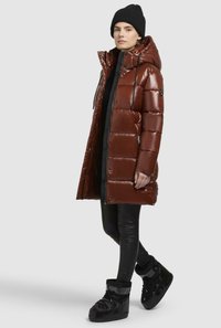 Bruine gewatteerde jas met glanzende afwerking, capuchon en zwarte accenten; gecombineerd met zwarte leggings en zwarte geïsoleerde laarzen. Kleding voor koud weer.