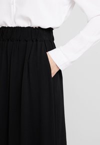 Gonna nera plissettata con vita elasticizzata, dotata di tasche laterali. Abbinata a una camicia bianca button-up, enfatizzando un look monocromatico.