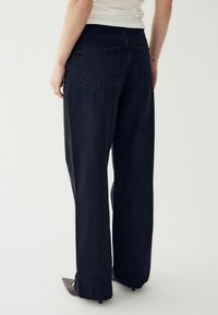 Aizmugures skats uz personu, kas valkā melnas platās bikses un tumšus norādītus apavus, stāvot pret vienkāršu baltu fonu.