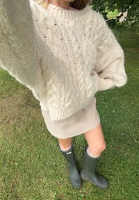 Fleckiger beiger Rock, grober Cream-Kabelstrickpulli und grüne Gummistiefel. Strukturiertes Material mit lässiger Passform und gemütlichem Design.