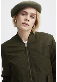 Veste bomber vert olive avec fermeture éclair, col et poignets côtelés. Assortie à un béret en feutre vert et des boucles d'oreilles créoles en argent.