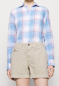 Osoba oblečená v béžových krátkých kalhotách na knoflíky a modré, bílo-červené kostkované košili s dlouhým rukávem a límečkem, stojící před jednobarevným pozadím.