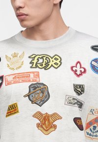 Jeune homme portant un sweat-shirt gris clair décoré de divers patchs brodés colorés et de logos.