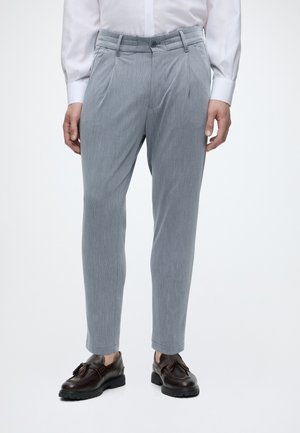 Persona che indossa pantaloni plissettati grigio chiaro, camicia bianca e mocassini marrone scuro con nappine, in piedi su uno sfondo semplice.