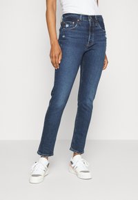 Jeans de mezclilla azul oscuro con ajuste ceñido, desgaste sutil, diseño de cinco bolsillos y costuras contrastantes. Combinados con zapatillas blancas.