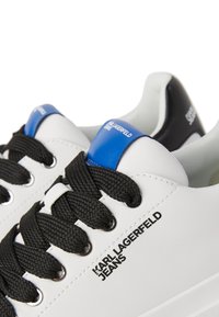 Adidași sintetici albi cu șireturi negre, având un accent albastru aproape de toc și cu "Karl Lagerfeld Jeans" imprimat pe lateral.