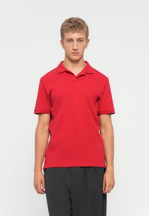 Young man with short blonde hair wearing a red short-sleeve polo shirt and dark grey pants standing against a plain white background.