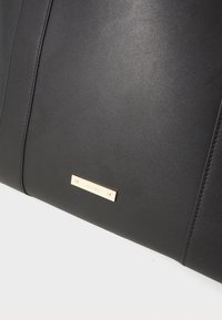 Sac en cuir noir au toucher lisse, avec une plaque logo métallique dorée et des accents de couture, forme rectangulaire pour un design structuré.