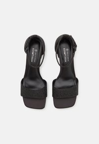Sandales à talons hauts véganes noires avec une bride de cheville, présentant une bandoulière texturée ornée de strass et une semelle intérieure lisse et mate.