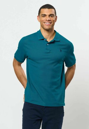 Homme souriant portant un polo à manches courtes bleu sarcelle et un pantalon foncé, debout avec les mains dans le dos, sur un fond uni.