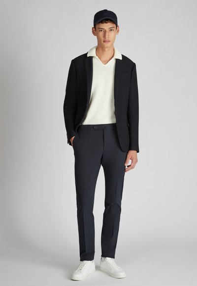 Blazer blu navy, maglione crema, pantaloni scuri slim-fit e sneakers bianche. L'outfit presenta un colletto e un design aderente. È indossato un cappellino nero.
