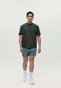 Moška temno zelena majica s kratkimi rokavi in napisom spredaj, v kombinaciji s svetlo modrimi kratkimi hlačami in belimi supergami. Gladko tkana tekstura.