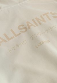 Béžová tkanina s hladkou textúrou, obsahujúca text "ALLSAINTS" a "LONDON" v embosovanom zlatom písme, spolu s japonským písmom.