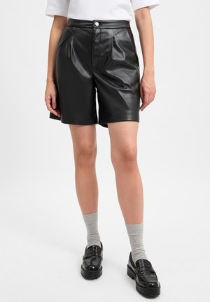 Black faux leather shorts with a high waist, pleats at the front, and a button closure. Styled with grey socks and chunky black loafers.
