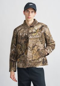 Καμουφλάζ μπουφάν σε καφέ και πράσινους τόνους, με μπροστινό κούμπωμα με κουμπιά, τσέπη στο στήθος και υφή που θυμίζει φλοιό και φύλλωμα.