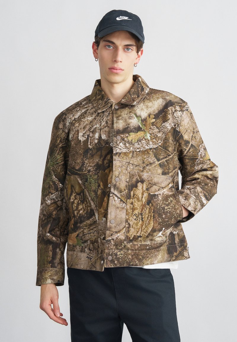 Καμουφλάζ μπουφάν σε καφέ και πράσινους τόνους, με μπροστινό κούμπωμα με κουμπιά, τσέπη στο στήθος και υφή που θυμίζει φλοιό και φύλλωμα.