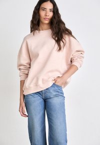Sudadera de color rosa claro, salpicada, con cuello redondo y hombros caídos, combinada con pantalones vaqueros de pierna ancha azules, que presentan un corte relajado.