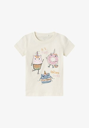 T-shirt color crema chiaro con una stampa grafica che raffigura tre dolcetti in stile cartone animato: un cupcake, una ciambella e una torta, con unicorni e la scritta "È magia."