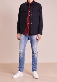 Giacca leggera navy con due tasche sul petto, indossata sopra una camicia a scacchi rossa, abbinata a jeans chiari strappati e sneakers bianche.