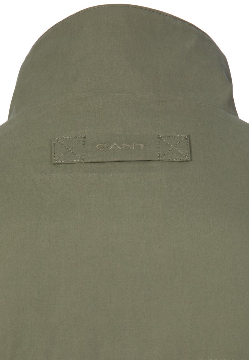 Colletto di giacca verde oliva con etichetta del marchio "GANT" cucita, posizionata al centro tra due toppe quadrate rinforzate.