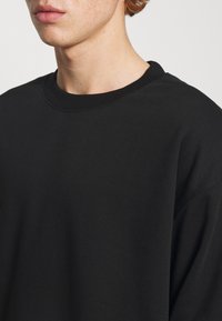 Sweatshirt noir en coton doux, avec un col rond et une coupe décontractée. Texture lisse avec des détails de couture visibles.