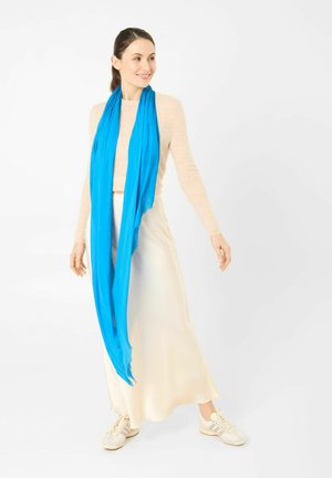 Frau trägt beige langärmliges Oberteil, cremefarbenen Rock, weiße Sneaker und einen leuchtend blauen Schal, steht vor weißem Hintergrund, lächelt und schaut seitlich.