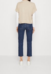 Top lavorato a maglia beige con maniche corte, abbinato a jeans dritti blu scuro e scarpe da ginnastica bianche. Tasche posteriori visibili sui jeans.