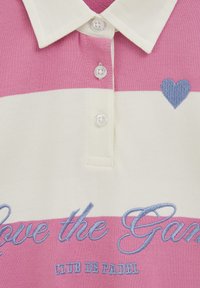 Poloshirt met roze en witte strepen, voorzien van een blauw geborduurd hart, drie knopen en de tekst "Love the Game" in blauw stiksel.