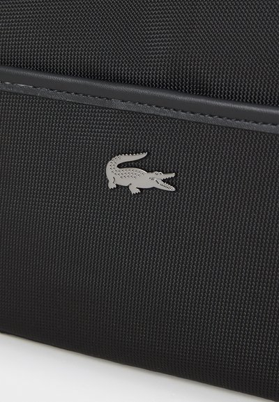 Sac en tissu noir texturé avec un logo de crocodile en métal argenté à l'avant et une finition en cuir lisse sur le dessus.