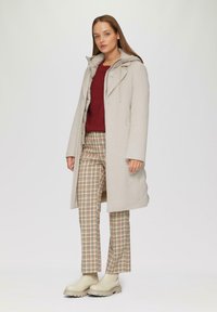 Beige wollen jas met een capuchon, drukknoopsluiting en lange mouwen, gecombineerd met een beige en bruine geruite broek en beige chunky laarzen.