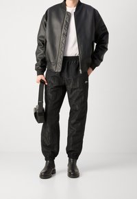 Černá bomber bunda s prošívaným designem, spárovaná s texturovanými černými tepláky. Černé boty a taška s sluchátky dokončují outfit.