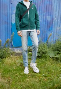 Green zip-up hoodie, white shirt, light blue jeans, black and white striped sneakers, standing on grass with blue wall in background.