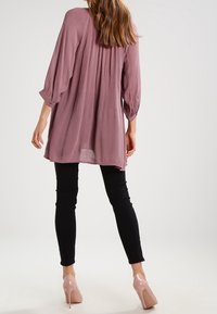 Letvægts, mauve tunika med trekvartærmer, plisseret rygdetalje, parret med sorte leggings og nude højhælede sko.