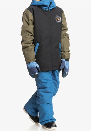 Enfant portant un pantalon de neige bleu, une veste noire et olive avec des poches bleues, des gants bleus et des bottes de neige noires, debout sur un fond blanc.