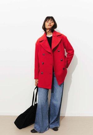 Femme portant un manteau rouge vif à double boutonnage, un jean bleu large, des chaussures noires à clous, tenant un grand sac noir sur fond blanc.