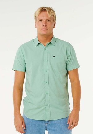 Blondyn stojący mężczyzna w zielonej koszuli na krótkim rękawie z guzikami oraz niebieskich dżinsach na jednolitym białym tle.