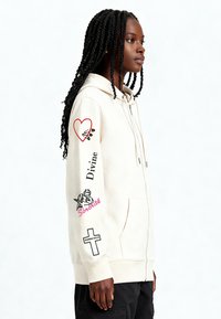 Kremowa bluza z kapturem zapinana na zamek z przodu, dwie kieszenie oraz nadruki graficzne na obu rękawach, w tym serca i napis "Divine". Miękka w dotyku.