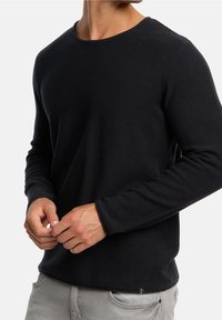 Chemise en tricot noire à manches longues avec un col rond, présentant un tissu texturé et des manches ajustées. Pantalons gris visibles à la taille.