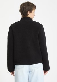 Giacca in pile nera con un esterno texturizzato, colletto alto e maniche lunghe. La vista posteriore mostra i dettagli delle cuciture e una vestibilità comoda.