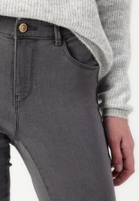 Pantalones de mezclilla gris de corte slim, con bolsillos frontales y un botón de bronce en la cinturilla. Se acompaña de un suéter de canalé gris claro.